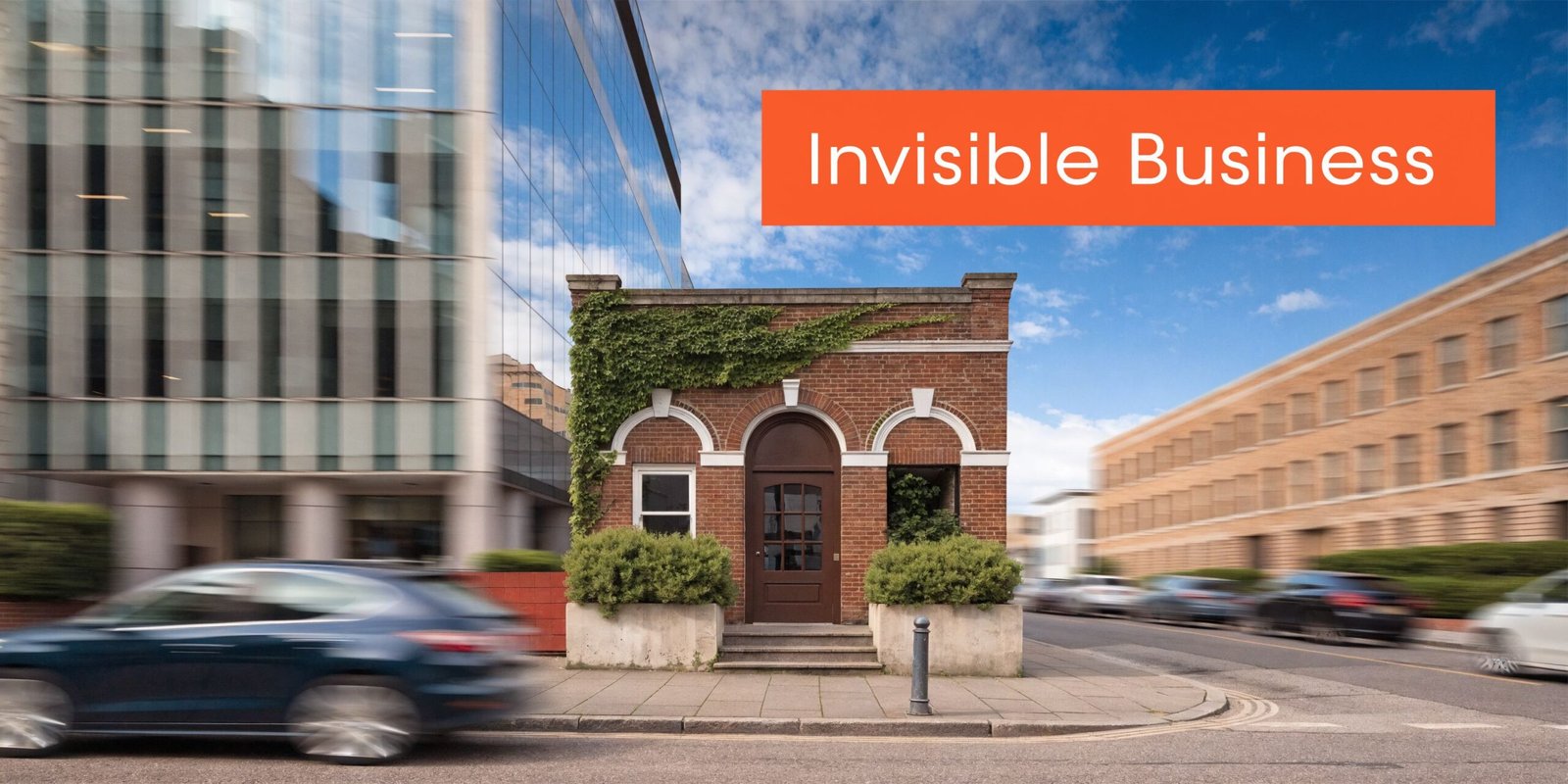 A small historic brick building standing between large modern office buildings with motion blurred cars passing by.